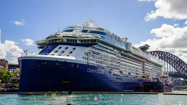Large Celebrity EDGE cruise ship docked near Sydney Harbour Bridge under blue sky with clouds.