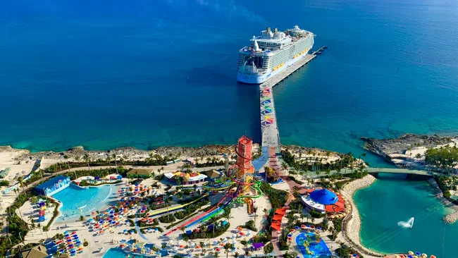 Aerial view of a colorful waterpark with pools, slides, umbrellas, and a large cruise ship docked nearby.