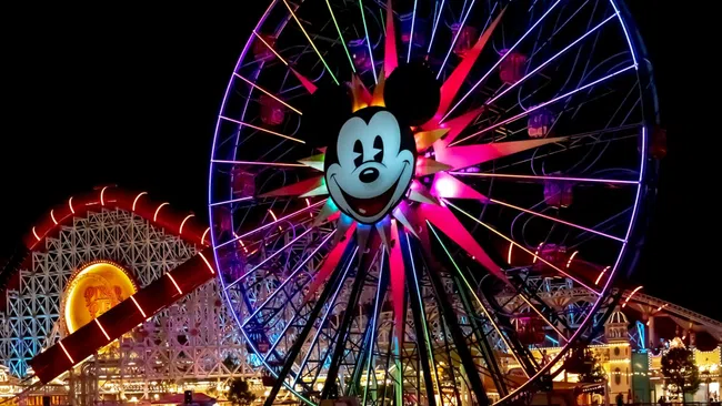 Night view of a brightly lit Mickey Mouse Ferris wheel and roller coaster with colorful reflections on water.