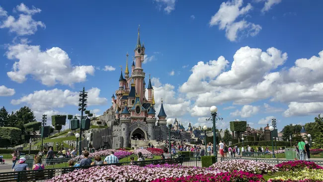 Disney castle surrounded by colorful flowers and visitors under a bright blue sky with fluffy clouds