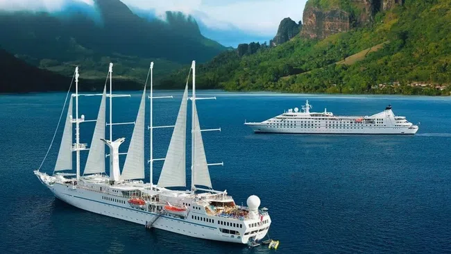 White sailing ship and cruise ship anchored in clear blue water near green mountainous island under cloudy sky.