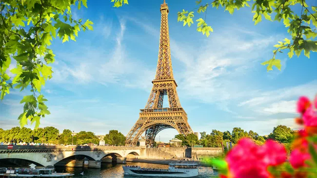 Bright Eiffel Tower by the Seine River with boats, framed by green leaves and pink flowers on a sunny day.