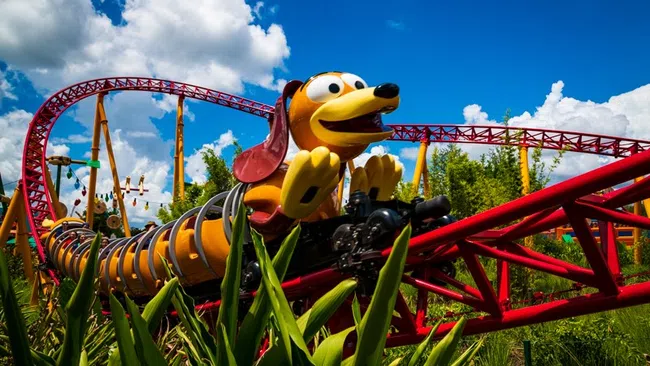 Roller coaster with a colorful cartoon dog train passing through green plants under a blue cloudy sky.