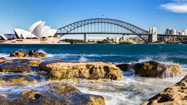 Sydney Opera House and Harbour Bridge with blue sky and rocky shoreline in foreground