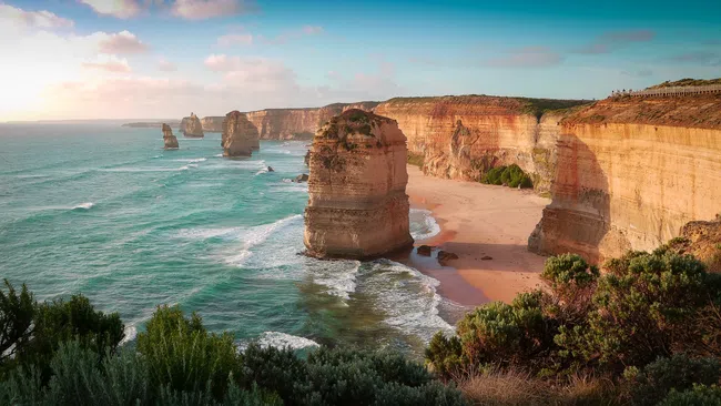 View of Twelve Apostles limestone stacks along the Great Ocean Road with cliffs, ocean waves, and sandy beach under a partly cloudy sky.