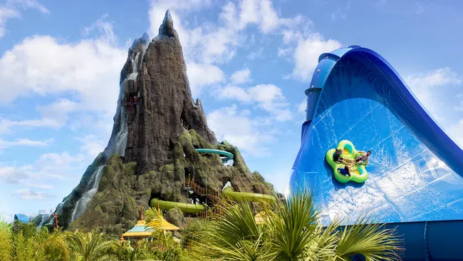 Thrill riders slide down vibrant blue water slide next to towering volcanic mountain in sunny water park.