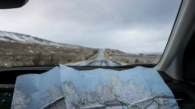 A folded map on a dashboard overlooking an empty road surrounded by mountains.
