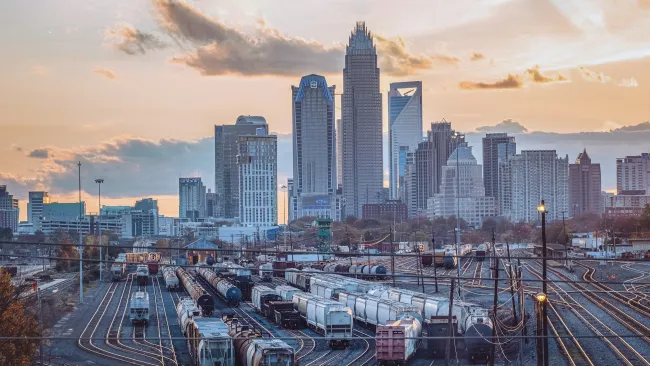 Urban train yard with multiple tracks and freight trains set against city skyline at sunset under cloudy sky