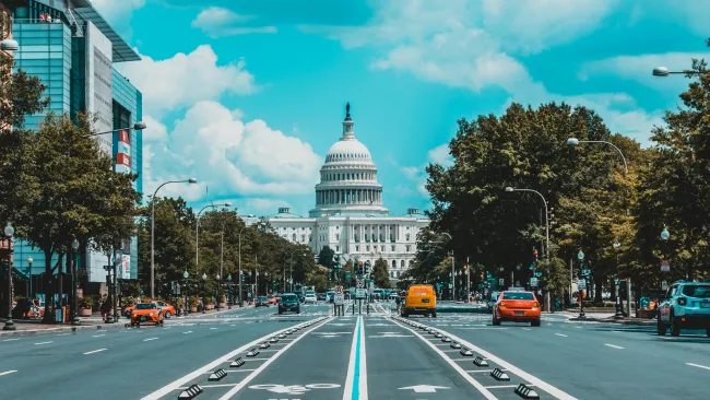 Wide street leading to the United States Capitol with bike lanes and cars under a partly cloudy sky.
