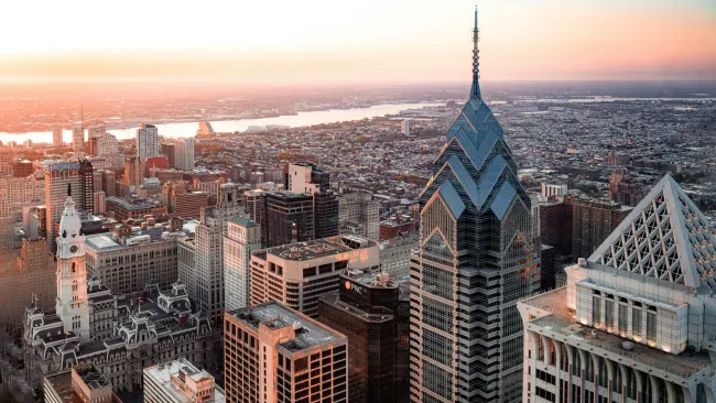 Aerial view of Philadelphia skyline at sunset with modern skyscrapers and historic buildings along a river.