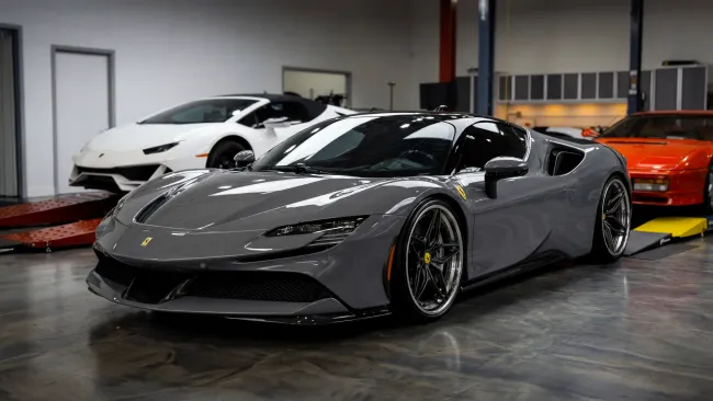Gray Ferrari sports car parked indoors with white and red luxury cars in the background in a showroom or garage.