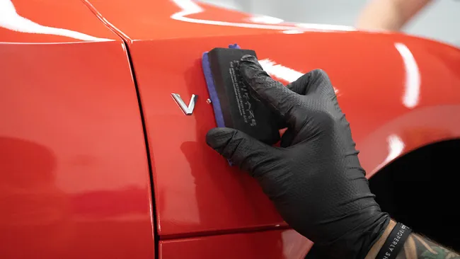 Hand wearing black glove applying polish on shiny red car door near silver V emblem