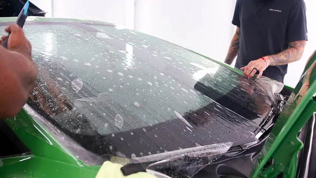 Two people applying protective film on a green car windshield with water bubbles visible