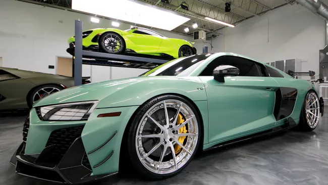 Matte green sports car with silver wheels and yellow brakes inside a modern garage with other luxury cars.
