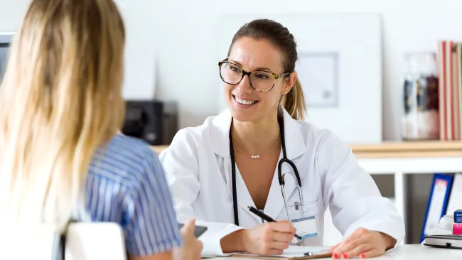 Smiling female doctor with glasses consulting a female patient in a bright medical office.
