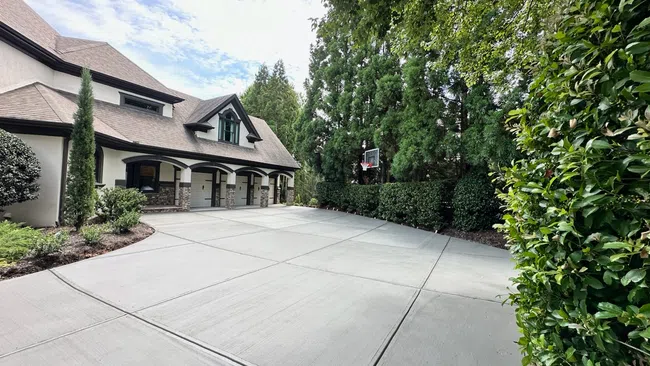 Spacious concrete driveway with basketball hoop beside a modern house surrounded by lush green trees and bushes.