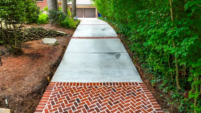 Concrete driveway with decorative brick inlays leading to a two-car garage surrounded by greenery and trees.