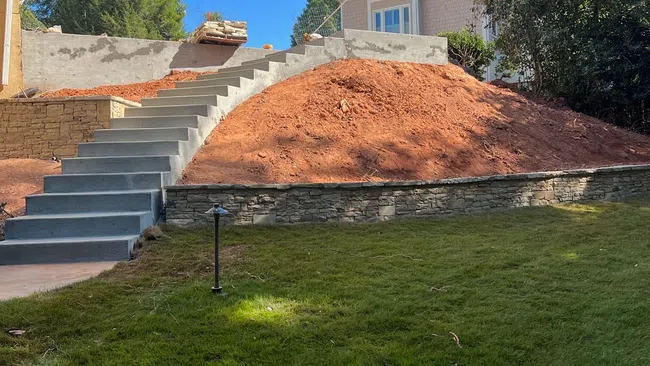 Concrete stairs lead up a landscaped hill with red soil, retaining wall, adjacent house, and green grass under blue sky.