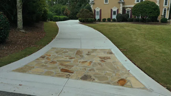 Curved driveway with a stone inlay leading to a beige house surrounded by green lawn and trees.