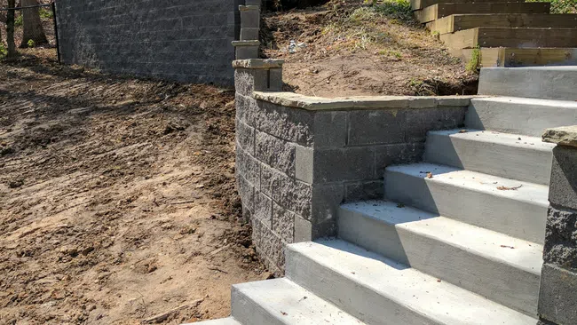 Newly constructed concrete stairs and retaining walls in a backyard with bare soil and trees.