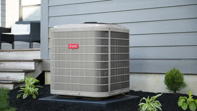 Outdoor Bryant air conditioning unit installed beside a gray house on a black mulch bed with plants around it.
