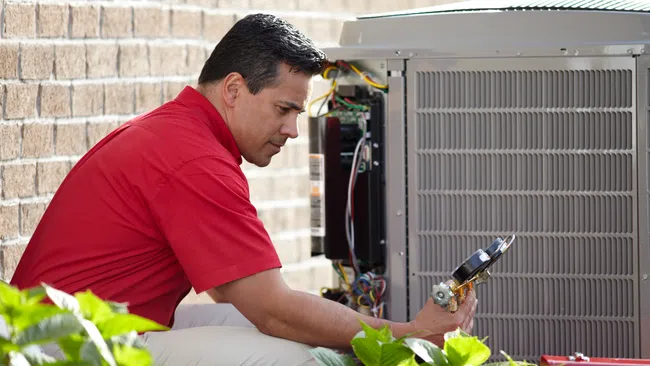 Technician in red shirt inspecting and repairing an outdoor air conditioning unit with tools near a brick wall.