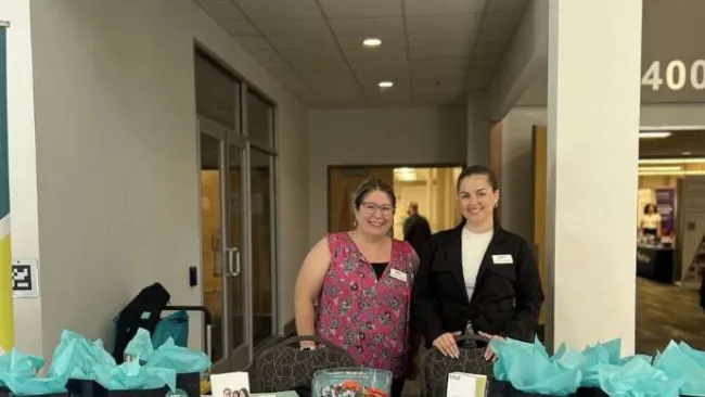 Two women at a promotional booth with branded gift bags, pens, and informational brochures at a conference