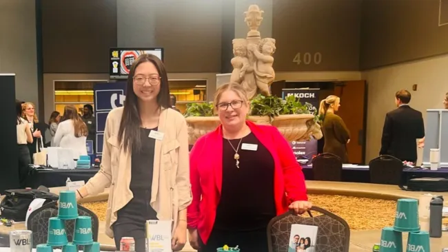 Two smiling women standing behind a promotional table with branded cups and flyers at an indoor event.