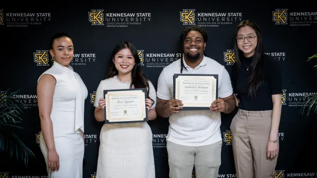 Four Kennesaw State University students stand in front of a branded backdrop, two holding School of Accountancy certificates.