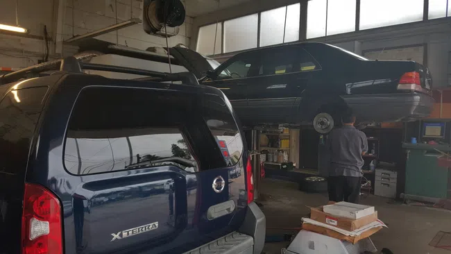 Blue Nissan Xterra and black sedan car in garage with mechanic inspecting vehicle on lift under bright windows.