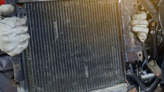Mechanic holding a dirty car radiator with gloved hands near an engine during repair work.