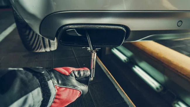 Mechanic wearing gloves uses a tool to work on a car's rear exhaust pipe in a workshop setting.