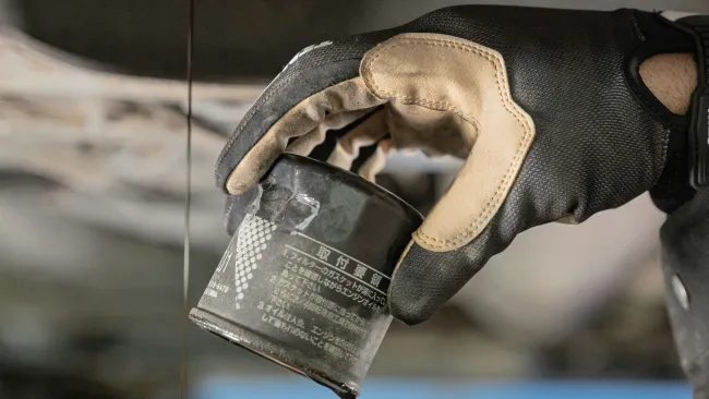 Gloved hand holding an oil filter while used engine oil drains from a vehicle undercarriage.
