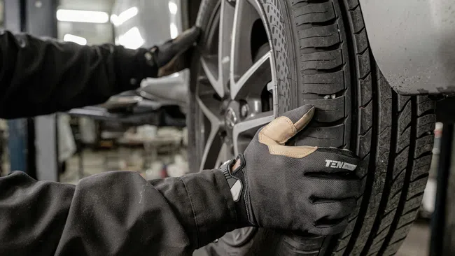 Mechanic wearing gloves inspecting a car tire in an auto repair shop with a lifted vehicle in the background.