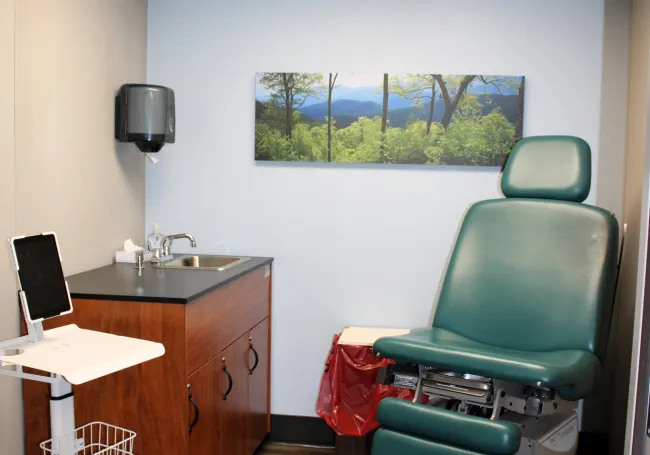 Clean medical exam room with green exam chair, wooden cabinets, sink, and nature artwork on wall..