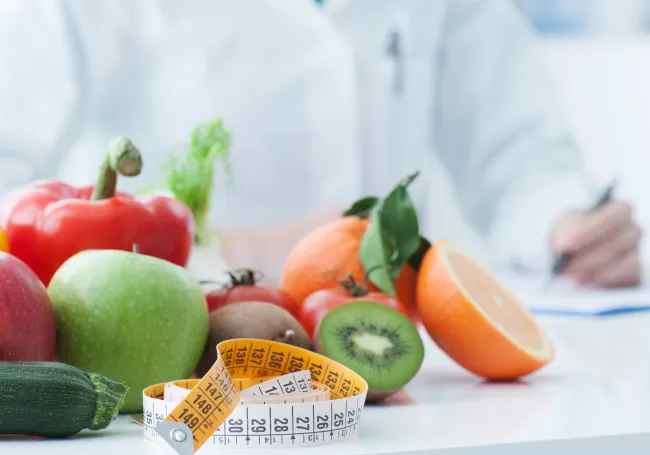 Fresh fruits and vegetables with a measuring tape in front of a person writing on a clipboard, symbolizing healthy eating..