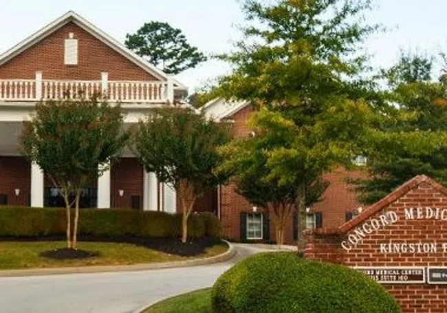 Brick medical park building with white columns and trees along curved driveway under cloudy sky.