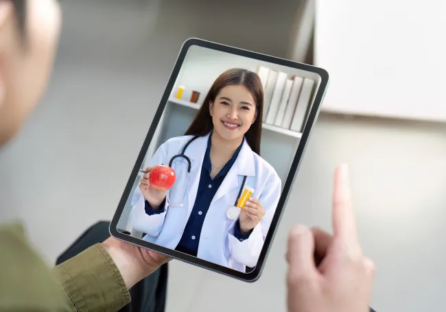 Patient video consulting with smiling female doctor holding an apple and medicine on a tablet screen.