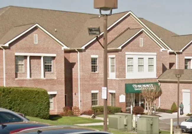 Brick commercial building with green awning and parking lot filled with cars on a clear day.