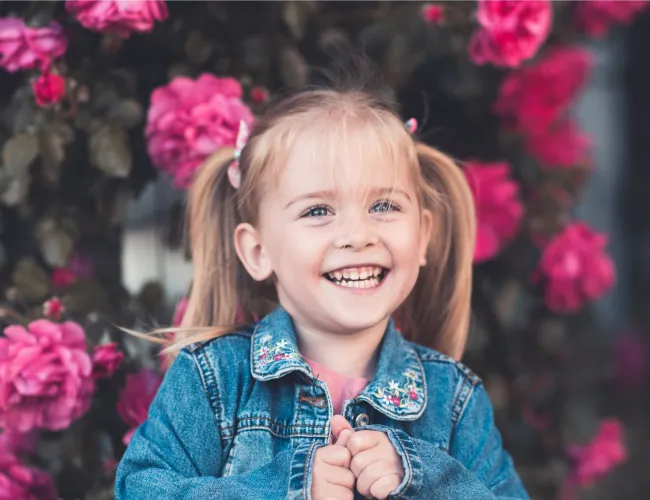 Smiling young girl with blonde hair in pigtails wearing a denim jacket standing against a backdrop of vibrant pink flowers