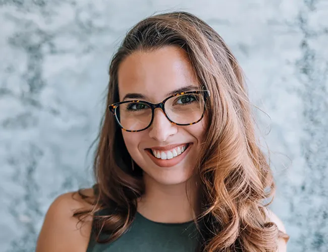 Smiling woman with long brown hair, wearing tortoiseshell glasses and a green sleeveless top, against a textured wall.