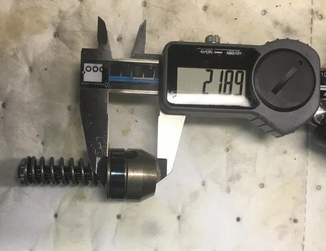 Digital caliper measuring a metal spring-loaded mechanical component on a stained white surface.