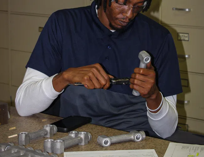 Technician inspecting metal parts with calipers on a table with documents and tools in an office setting.