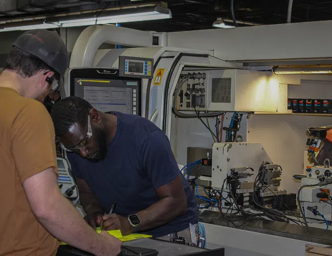 Two factory workers in safety glasses operate and inspect a CNC machine in an industrial manufacturing setting.