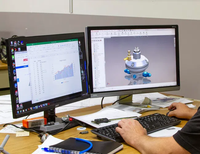 Man working on dual monitors with 3D engineering design and data charts, desk cluttered with papers and notebook.