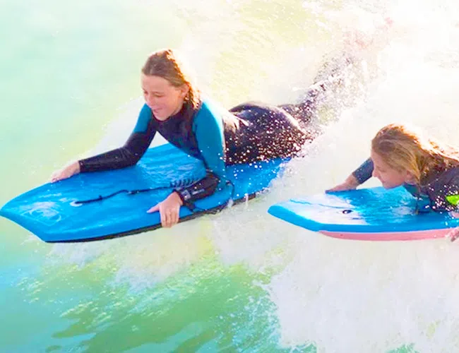 Two girls wearing wetsuits riding bodyboards on ocean waves enjoying a sunny day at the beach.