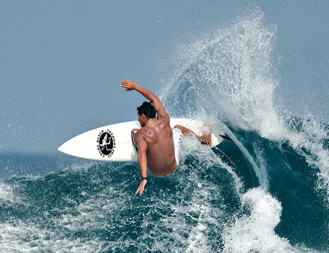 Surfer riding a powerful ocean wave, performing a sharp turn with water spray against a clear blue sky.