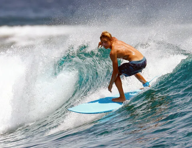 Surfer riding a blue surfboard on a breaking ocean wave under clear sky.