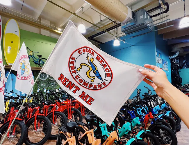 Hand holding a Cocoa Beach Surf Company flag in front of colorful rental bikes inside a shop with surfboards.