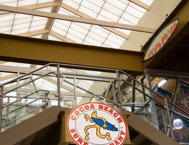 Cocoa Beach Surf Company logo sign hanging inside a modern building with glass railings and a skylight ceiling.